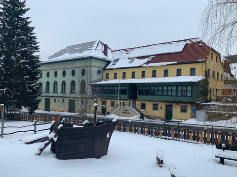 Ein verschneiter Spielplatz mit einem Holzschiff im Vordergrund, dahinter ein großes, gelb-grünes Gebäude mit roten Dächern und Tannenbaum.