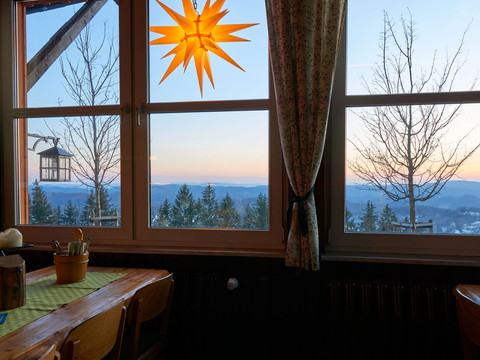 Bekijk Wachbergbaude Holztisch mit Kerze und Besteck vor einem Fenster mit Blick auf schneebedeckte Bäume und Berge bei Sonnenuntergang; ein leuchtender Stern hängt im Fenster.
