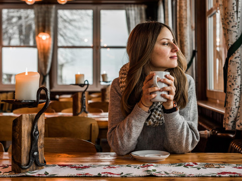 Gastenkamer Wachbergbaude Eine Frau in einem gemütlichen Café hält eine Tasse, schaut aus dem Fenster; auf dem Tisch brennt eine Kerze, warme Atmosphäre.