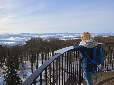 Uitzicht vanaf de Prinz Georg toren op de Ungerberg Frau mit blauem Rucksack und grauem Schal steht auf einem Aussichtspunkt und blickt über eine verschneite Landschaft mit Bäumen und Hügeln.