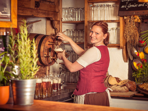 "Zum Musketier" Restoration Königstein Fortress Eine Frau in roter Weste zapft Bier aus einem Holzfass in einer rustikalen Bar; im Hintergrund sind Gläser und frisches Gemüse zu sehen.