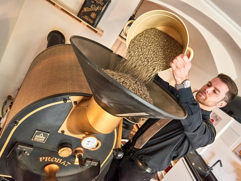 Café & Wijnhuis Koffiebranderij Schmole in Pirna Ein Mann in schwarzer Kleidung schüttet grüne Kaffeebohnen aus einem gelben Behälter in eine goldene Kaffeeröstmaschine.