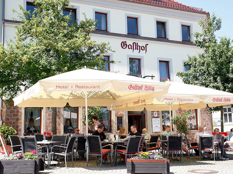 Außenbereich eines Gasthofs mit beige Sonnenschirmen und mehreren Menschen, die an Tischen sitzen; entspannte Atmosphäre bei sonnigem Wetter.