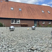 image00009.jpeg Ein rustikales Backsteingebäude mit rotem Ziegeldach im ländlichen Stil; im Vordergrund ein Boulespielfeld mit glänzenden Kugeln und einem kleinen Zielball.