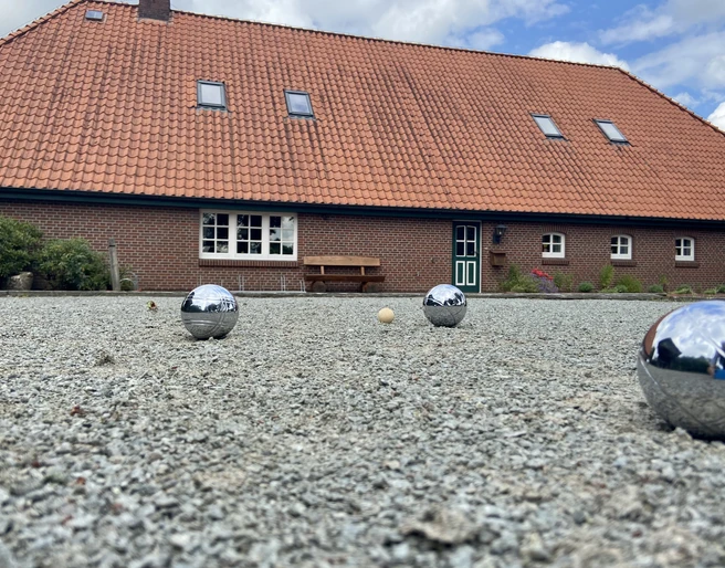 image00009.jpeg Ein rustikales Backsteingebäude mit rotem Ziegeldach im ländlichen Stil; im Vordergrund ein Boulespielfeld mit glänzenden Kugeln und einem kleinen Zielball.