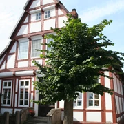 Fachwerk Hoya Fachwerkhaus in Hoya mit markanter weiß-roter Fassade und zwei Treppenstufen, umgeben von Bäumen.