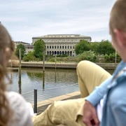 Stadthalle, Mülheim an der Ruhr Blick auf die Stadthalle Mülheim, ein imposantes Gebäude am Fluss mit Bäumen und Menschen im Vordergrund.