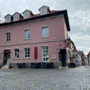 Pup Dat Wählige Rott Historisches rosafarbenes Gebäude mit Hotel, an einer gepflasterten Straße gelegen, mehrere Fenster und Dachgauben.