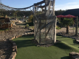 Turm Activity Park Hagenburg Der Turm im Vordergrund eines Minigolfplatzes ist von Netzen umgeben, im Hintergrund sind Pflanzen.