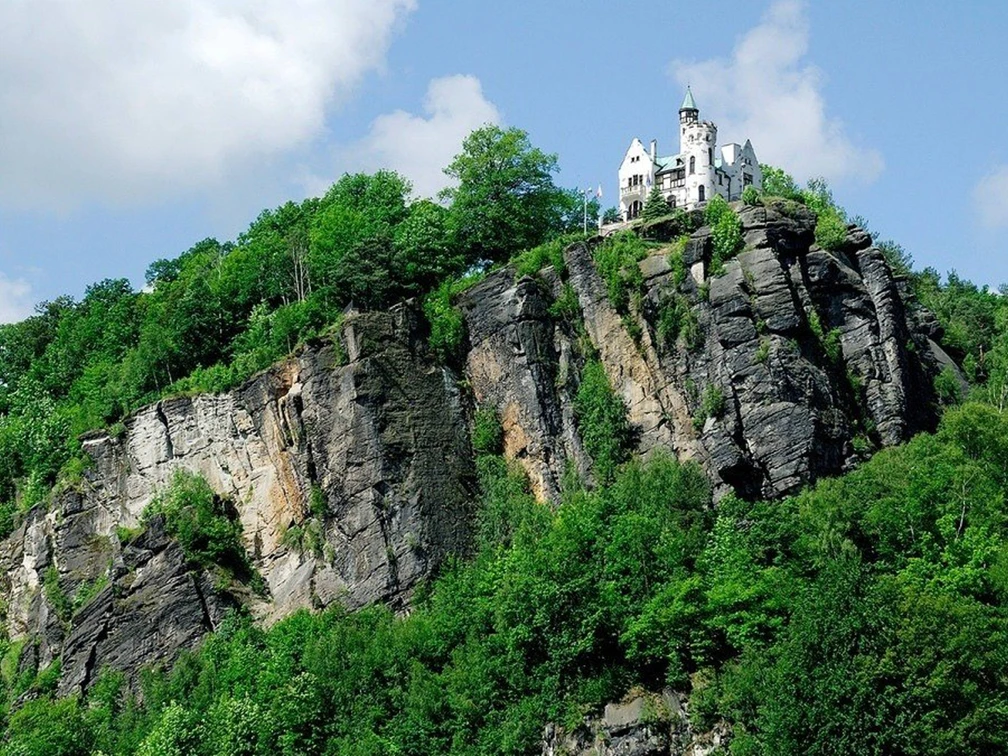 Klettersteig Děčín Ein weißes Schloss thront auf einem bewaldeten Felsen unter blauem Himmel mit Wolken, umgeben von üppigem Grün.