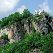 Klettersteig Děčín Ein weißes Schloss thront auf einem bewaldeten Felsen unter blauem Himmel mit Wolken, umgeben von üppigem Grün.