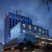 Modernes Hotelgebäude mit blauer Glasfassade und beleuchtetem Dach bei bewölktem Abendhimmel.
