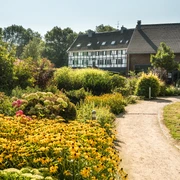 Hofcafé Abtsküche in Heiligenhaus Ein Weg führt durch einen üppig bepflanzten Garten zu einem Fachwerkhaus unter klarem Himmel.