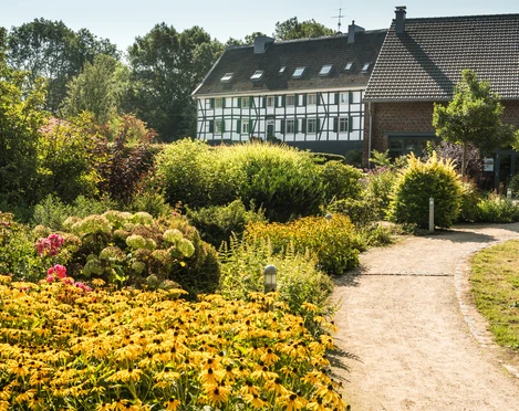 Hofcafé Abtsküche in Heiligenhaus Ein Weg führt durch einen üppig bepflanzten Garten zu einem Fachwerkhaus unter klarem Himmel.
