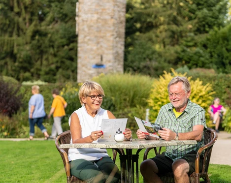 am Hofcafé Abtsküche in Heiligenhaus Ein älteres Paar liest entspannt an einem Tisch im grünen Garten vor einem steinernen Turm.