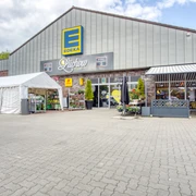Außenansicht Edeka Lüchow  Die Außenansicht des Edeka-Marktes in Lüchow zeigt einen weitläufigen Parkplatz vor einem modernen Ladengebäude. Das flache Dach und die großen Glasfenster prägen das Erscheinungsbild.