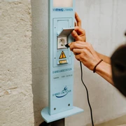 E-Bike Aufladestation Eine Person schließt ein Ladekabel an eine E-Bike-Ladestation an einer hellen Wand an.