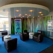 Eine moderne Rezeption mit Ledersesseln um kleine Tische auf blauem Teppichkreis, umgeben von Prospekten.