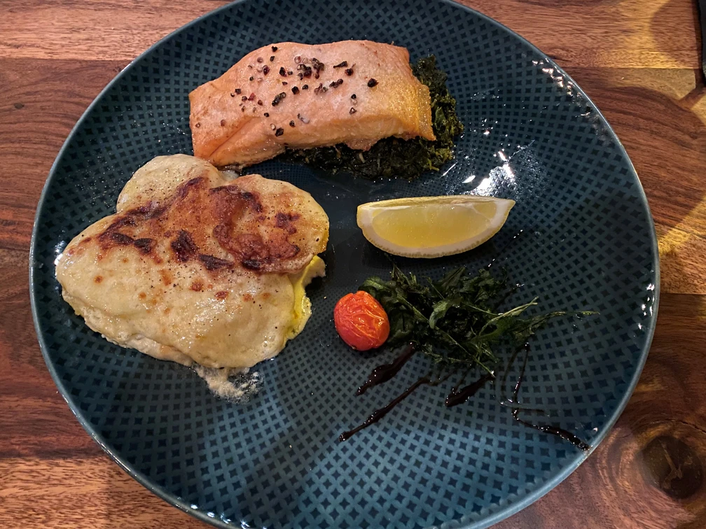 Lachsgericht Lachsfilet auf einem Bett aus grünem Gemüse mit Kartoffelgratin, Kirschtomate und Zitronenspalte.