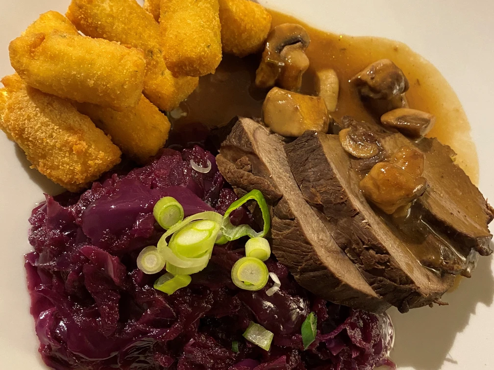 Hirschbraten Ein Teller mit Hirschbraten, Rotkohl, Kroketten und Pilzsoße, garniert mit Frühlingszwiebeln.