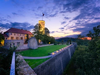 Bielefeld-Sparrenburg-blau-Wienke_Baustein und Teaser.jpg Burg mit rundem Turm und dahinter idyllischem Sonnenuntergang, umgeben von üppigem Grün und Mauer.
