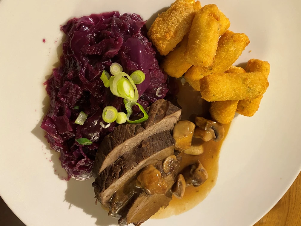 Hirschbraten Hirschbraten mit glasierten Pilzen, serviert mit Rotkohl und knusprigen Kroketten auf weißem Teller.