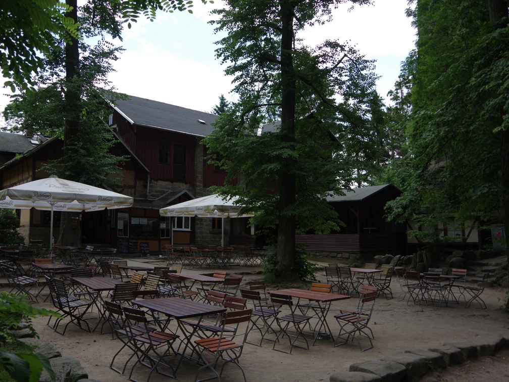 Berggaststätte Pfaffenstein Ein leerer Biergarten mit Holztischen und -stühlen unter großen Bäumen und Sonnenschirmen vor einem rustikalen Gebäude.