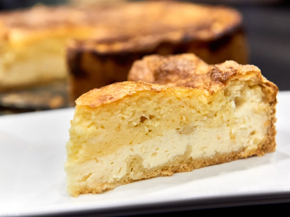 Wie macht man eine Eierschecke - Der typische Kuchen aus der Sächsischen Schweiz Ein Stück goldbrauner Käsekuchen auf einem weißen Teller, im Hintergrund der restliche Kuchen.
