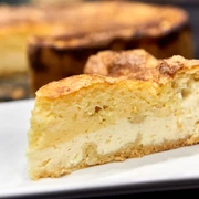 Wie macht man eine Eierschecke - Der typische Kuchen aus der Sächsischen Schweiz Ein Stück goldbrauner Käsekuchen auf einem weißen Teller, im Hintergrund der restliche Kuchen.