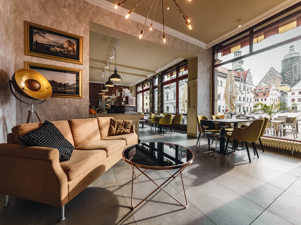 Restaurant Canaletto Gemütliches Café mit beiger Couch, schwarzem Kissen, rundem Glastisch und großen Fenstern mit Blick auf historische Gebäude.