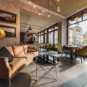 Restaurant Canaletto Gemütliches Café mit beiger Couch, schwarzem Kissen, rundem Glastisch und großen Fenstern mit Blick auf historische Gebäude.
