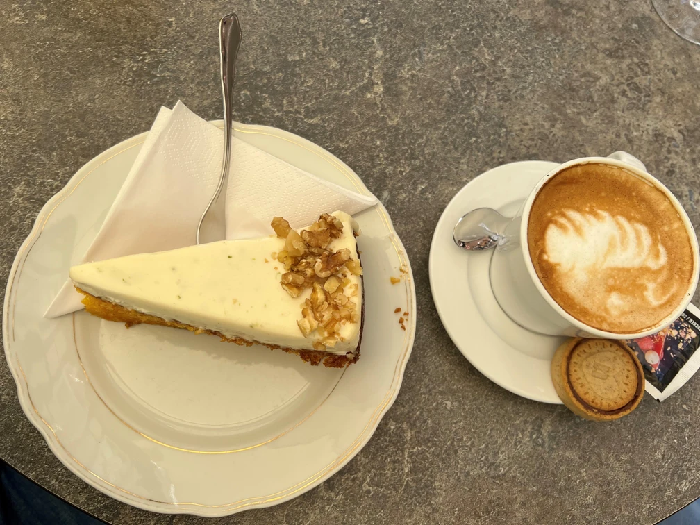 Ein Stück Walnusskuchen neben einer Tasse Cappuccino mit Keks auf einem steinernen Tisch serviert.