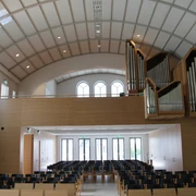 Moderner Kirchenraum mit geschwungenem Dach, Orgel links, hellem Holz, leeren Stuhlreihen und Fenstern.