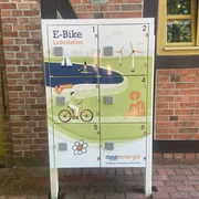 Ladestation für E-Bikes mit sechs Schließfächern vor roter Backsteinwand mit Holzrahmenfenster.
