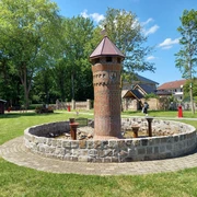 Mehrgenerationenplatz "Lüttje Festung" in Apen Der Mehrgenerationenplatz "Lüttje Festung" in Apen zeigt einen zentralen Backsteinturm inmitten einer kreisförmigen Steinmauer, umgeben von gepflegtem Rasen und Bäumen.