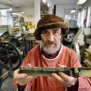 Die Buchmacherey in Velbert-Langenberg mit Hans Josef Altmann.jpg Ein Buchdrucker in traditioneller Kleidung hält eine Druckplatte in einer historischen Werkstatt.