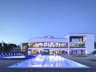 Aussenbereich der carpesol SPA Therme Moderne Gebäude mit beleuchtetem Pool und gläserner Kuppel vor Abendhimmel.