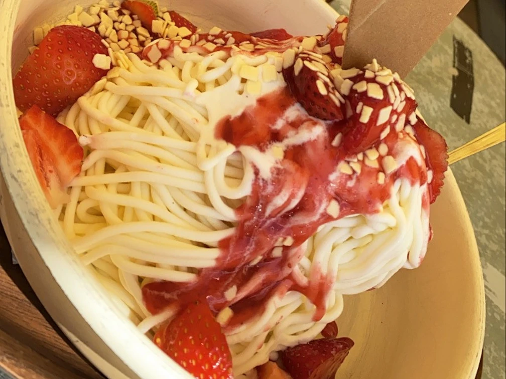 Eis Spaghetti-Eis mit Erdbeeren, Sahne, roter Erdbeersoße und Schokoraspeln in einer weißen Schale.