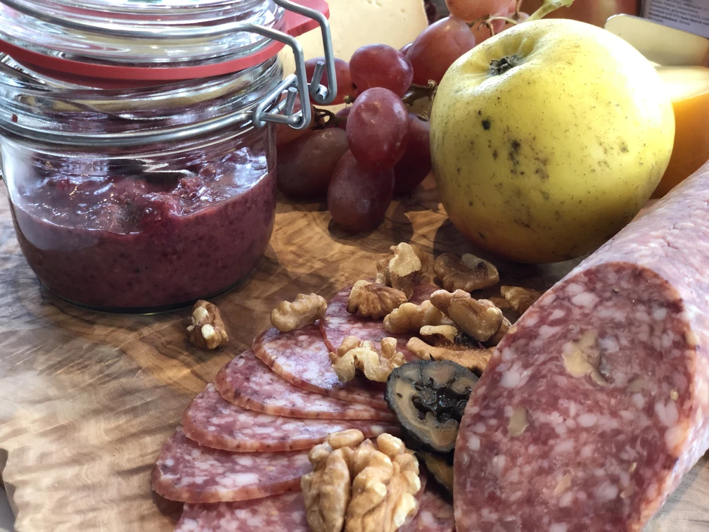 Kalte Platte Verschiedene Wurstsorten, Nüsse und Trauben liegen auf einem Holzbrett neben einem Glas Aufstrich.