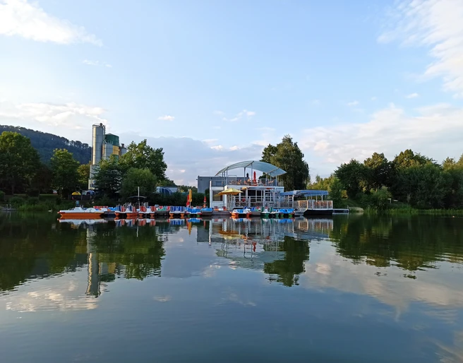 Wasserinn_freizeitgelaene_ahlemeyer.jpg Freizeitgelände mit Tretbootanleger und Café am ruhigen See unter blauem Himmel und Wolken.