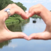 Aalschokker Weser Hände formen Herz und rahmen einen traditionellen Aalschokker auf einem ruhigen Fluss ein.