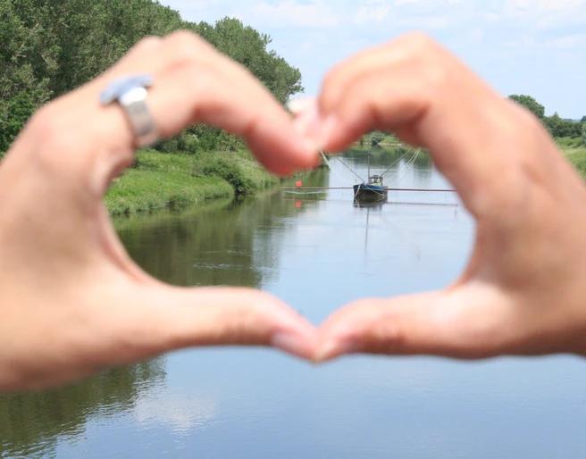 Aalschokker Weser Hände formen Herz und rahmen einen traditionellen Aalschokker auf einem ruhigen Fluss ein.
