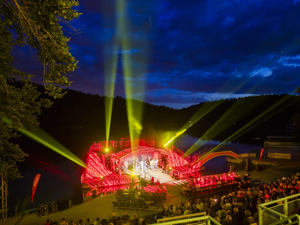 Seebühne Kriebstein - Kultur in Leipzig und Region Der Blick von den oberen Zuschauerrängen auf die Seebühne Kriebstein, ein Konzert wird gespielt, die Zuschauerplätze sind gefüllt und die Scheinwerfer strahlen in die verschiedenen Himmelsrichtungen zur blauen Stunde, Kultur, Veranstaltung
