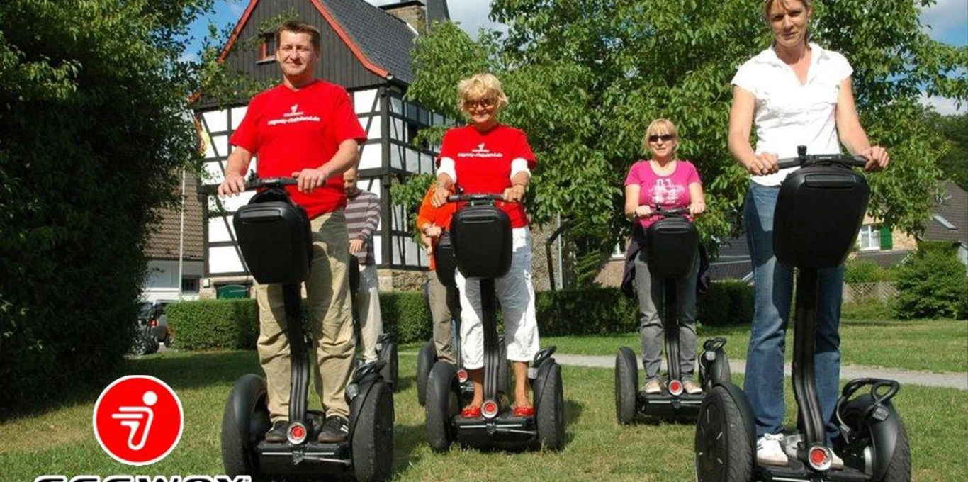 SEGWAYPoint-Haan-Rheinland Vier Personen fahren fröhlich auf Segways entlang eines grünen Wegs vor einem Fachwerkhaus.