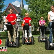 SEGWAYPoint-Haan-Rheinland Vier Personen fahren fröhlich auf Segways entlang eines grünen Wegs vor einem Fachwerkhaus.