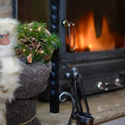 Eine Weihnachtsfigur mit Brille und grauem Mantel hält einen kleinen Tannenzweig neben einem brennenden Kamin; gemütliche Atmosphäre.