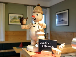 Schneemann im Gasthaus Zirkelstein Holzfigur eines lächelnden Schneemanns mit Vogel und Laterne, daneben ein Schild mit "Herzlich willkommen" in einem gemütlichen Raum.