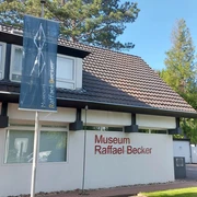 Museum Raffael Becker Das Bild zeigt die helle Fassade des modernen Museums Raffael Becker. Vorne ein Parkplatz mit Auto. Rechteckige Fenster und klare Linien prägen die Architektur, während umgebender, dichter Baumgrün dem Gebäude Leben einhaucht.The picture shows the bright façade of the modern Raffael Becker Museum. In front is a parking lot with a car. Rectangular windows and clear lines characterize the architecture, while the surrounding dense greenery breathes life into the building.