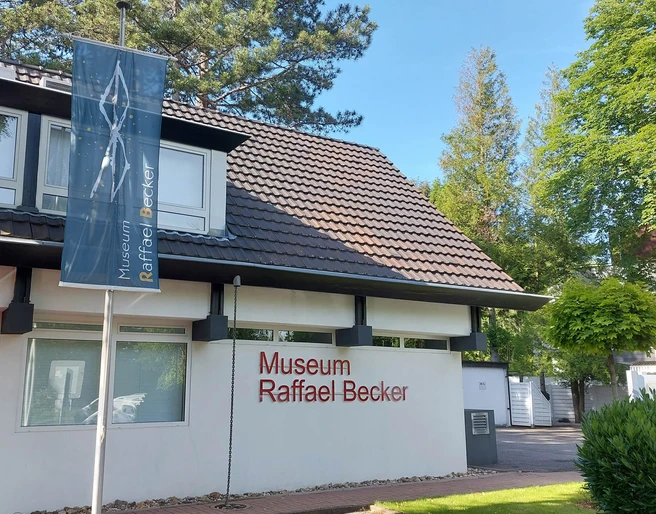 Museum Raffael Becker Das Bild zeigt die helle Fassade des modernen Museums Raffael Becker. Vorne ein Parkplatz mit Auto. Rechteckige Fenster und klare Linien prägen die Architektur, während umgebender, dichter Baumgrün dem Gebäude Leben einhaucht.The picture shows the bright façade of the modern Raffael Becker Museum. In front is a parking lot with a car. Rectangular windows and clear lines characterize the architecture, while the surrounding dense greenery breathes life into the building.