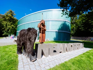 Neanderthal-Museum-MR-N-Tinka-Neandertal-Schriftzug-Mettmann.jpg Neanderthal-Museum in Mettmann mit einer Skulptur von Neandertaler und einem Wollnashorn im Freien.